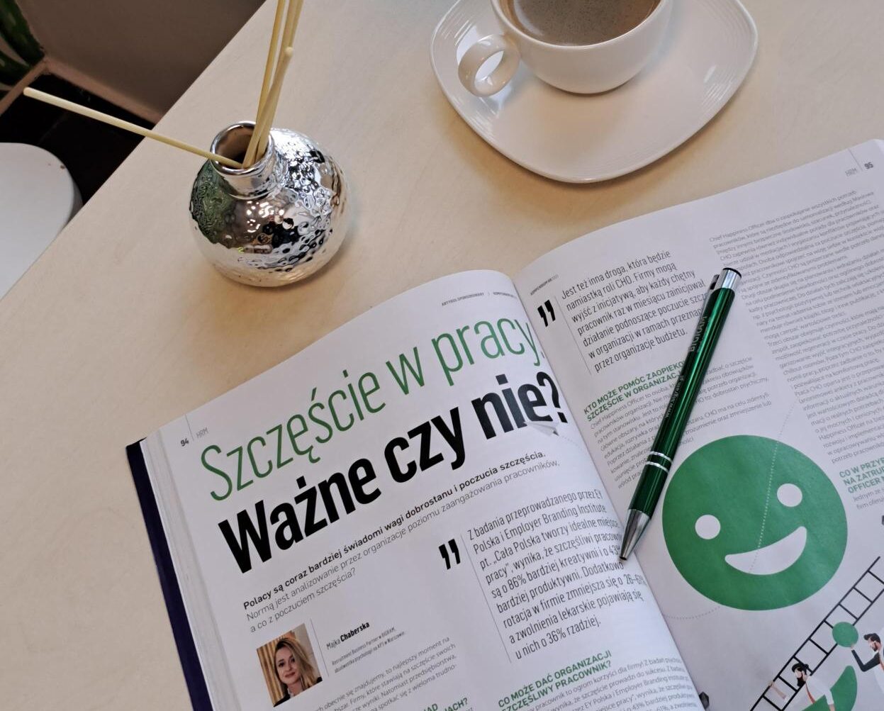 Otwarte czasopismo z artykułem o szczęściu w pracy leży na stole, obok stoi filiżanka kawy.